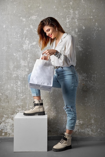 Primo piano della ragazza dei pantaloni a vita bassa che indossa blue jeans e tiene in mano il modello di pacchetto di carta artigianale in bianco del pacchetto bianco del modello con le maniglie