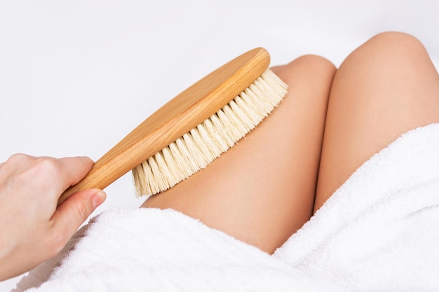 Primo piano delle cosce di una giovane donna magra con un asciugamano bianco che massaggia il suo corpo con una spazzola di legno dura