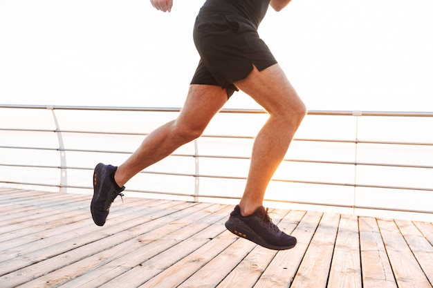 Primo piano delle gambe del giovane attivo in tuta sportiva che corre lungo il pilastro o il sentiero costiero in riva al mare, nella mattina