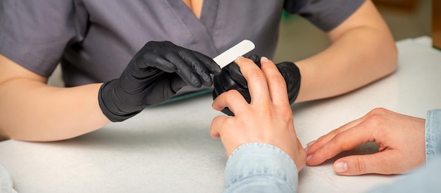 Primo piano delle mani caucasiche di un manicure professionista stanno limando le unghie di una giovane donna Giovane donna caucasica che riceve una manicure da un'estetista con una lima per unghie in un salone di bellezza