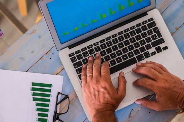 Primo piano delle mani che scrivono e lavorano con il computer portatile in ufficio su un tavolo di legno blu con statistiche e grafici