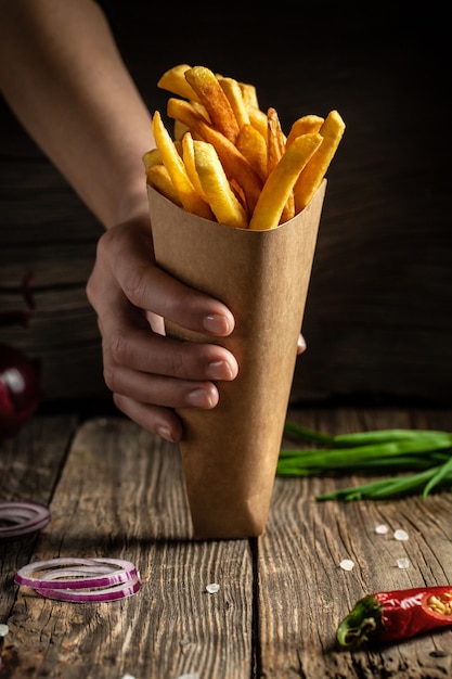 Primo piano delle mani con gustose patatine fritte al forno fatte in casa in scatola di carta sul tavolo di legno