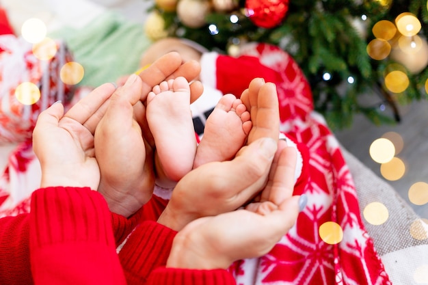 Primo piano delle mani dei genitori di mamma e papà che tengono i piedi del bambino all'albero di Natale con un decoro a forma di cuore rosso, il concetto di capodanno e Natale e una giovane famiglia felice