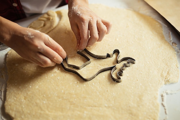 Primo piano delle mani del bambino che tagliano i biscotti dall'impasto. Ragazzo in grembiule che preme forma dell'alce della taglierina del biscotto. cuocere i biscotti di natale nella cucina di casa. Passatempo