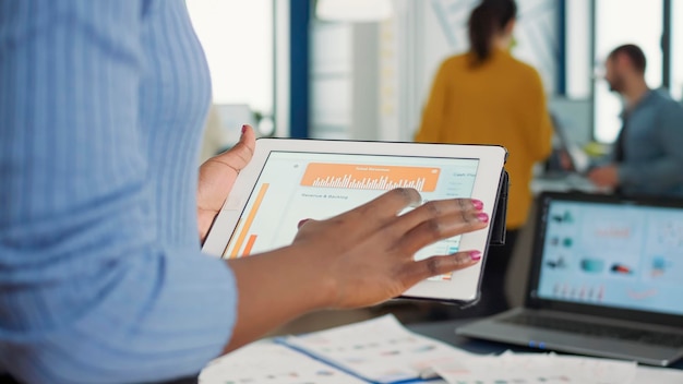 Primo piano delle mani della donna afroamericana che raccolgono il tablet che interagisce con il touchscreen guardando l'interfaccia del software erp in piedi alla scrivania. Messa a fuoco selettiva sul dipendente di avvio in possesso di dispositivo digitale.