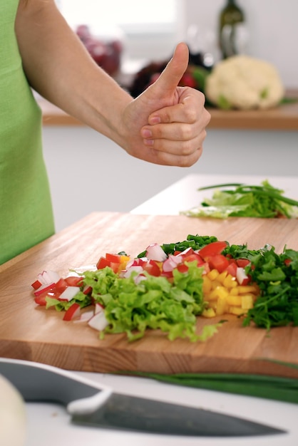 Primo piano delle mani della donna che cucinano in cucina. Casalinga che offre insalata fresca con i pollici in su. Concetto di cucina vegetariana e sana.