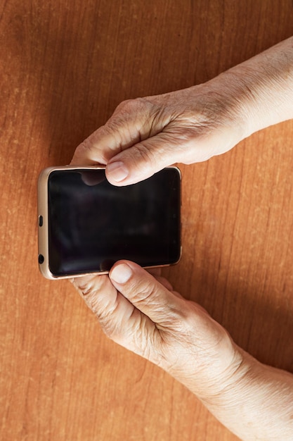 Primo piano delle mani della donna senior che tengono il telefono cellulare sul tavolo di legno