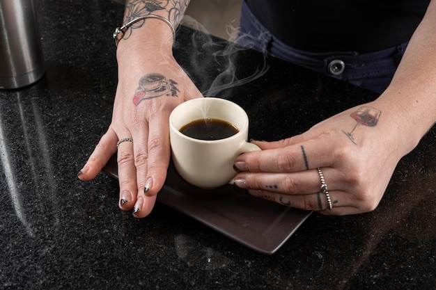 Primo piano delle mani della donna tatuata che tengono una tazza di caffè con fumo a forma di cuore