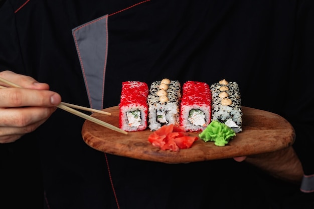 Primo piano delle mani di un cuoco unico in uniforme nera che tiene un vassoio di legno piatto pieno di sushi