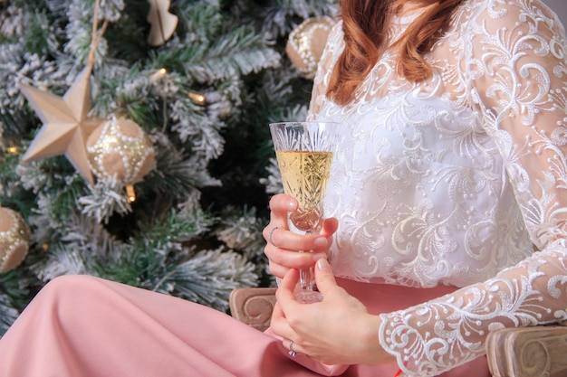 Primo piano delle mani di una donna in abito festivo che tiene un bicchiere di champagne vicino all'abete decorato durante la festa di Natale o Capodanno