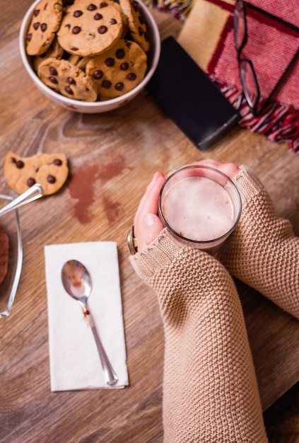 Primo piano delle mani femminili che tengono un bicchiere di bevanda calda su un tavolo con biscotti con scaglie di cioccolato su una ciotola di stelle