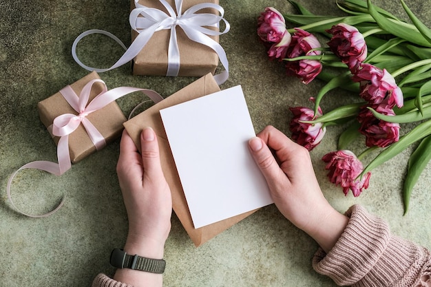 Primo piano delle mani femminili che tengono una busta con una cartolina di carta vuota sullo sfondo di un bouq