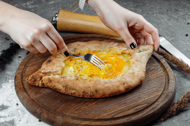 Primo piano delle mani femminili con un coltello e una forchetta mescolare il formaggio all'uovo in khachapuri adjarian su a