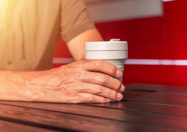 Primo piano delle mani maschili che tiene la tazza di caffè ecologica da asporto in silicone riutilizzabile nelle mani sopra il tabl di strada in legno...