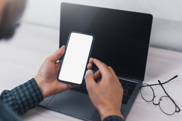 Primo piano delle mani maschili, tenendo lo smartphone sulla parete del computer portatile sulla scrivania.