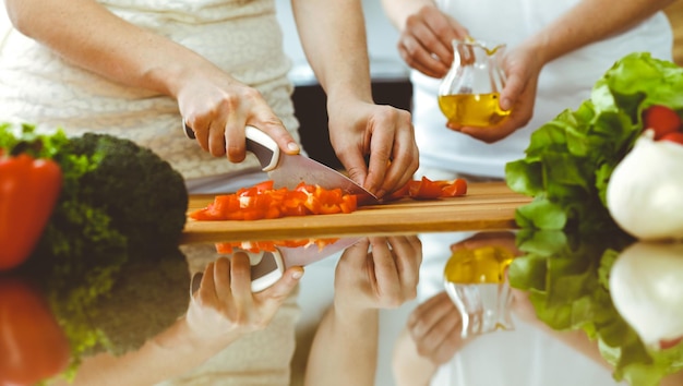 Primo piano delle mani umane che cucinano in cucina. Madre e figlia o due amiche che tagliano le verdure per insalata fresca. Amicizia, cena in famiglia e concetti di stile di vita.