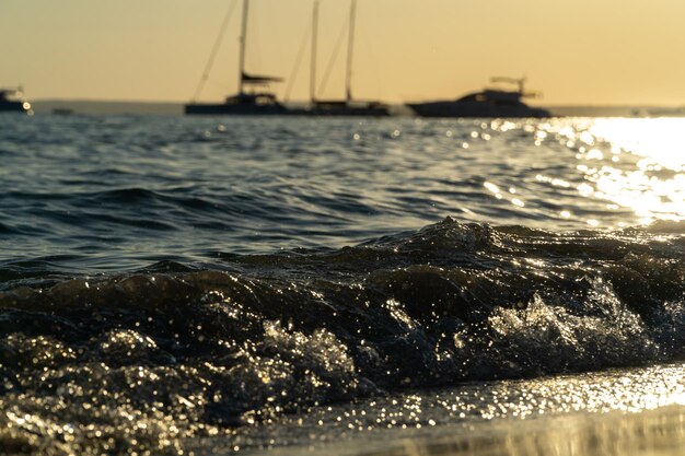 Primo piano delle onde del mare al tramonto con la silhouette di alcune barche sullo sfondo