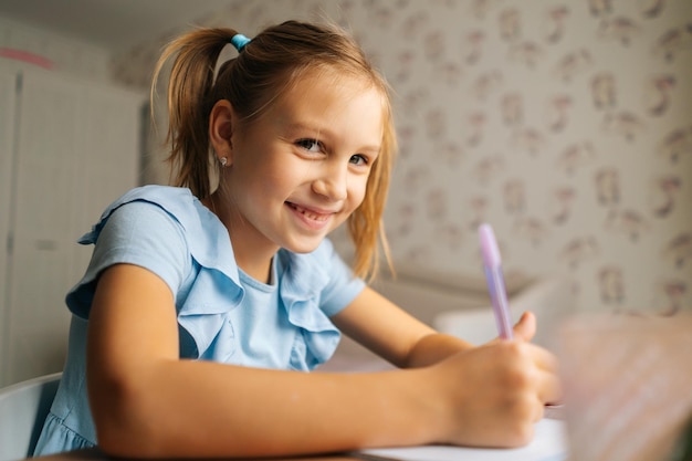 Primo piano di allegra bambina elementare che scrive facendo i compiti seduto a casa a tavola vicino