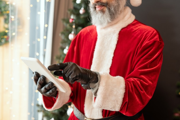 Primo piano di Babbo Natale barbuto in costume rosso e guanti di pelle con tablet in soggiorno