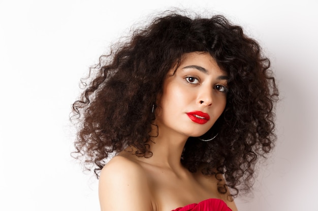 Primo piano di bella donna caucasica con taglio di capelli ricci e rossetto rosso, guardando tenero a porte chiuse, in piedi su sfondo bianco.