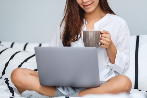 Primo piano di bella donna che utilizza e che lavora al computer portatile, bevendo caffè mentre sedendosi su un letto accogliente bianco a casa