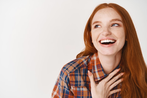 Primo piano di bella donna con i capelli rossi e la pelle pallida che ride felice, tenendo la mano sul cuore e guardando a sinistra il logo, in piedi sul muro bianco