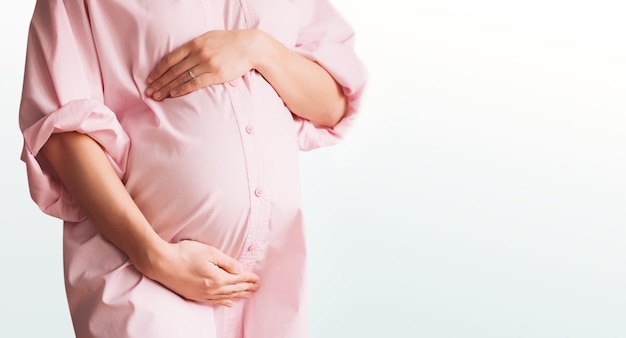 Primo piano di bella donna incinta in abito camicia rosa tiene le mani sul ventre su sfondo bianco Madre amorevole in attesa della nascita del bambino Preparazione alla gravidanza e concetto di aspettativa