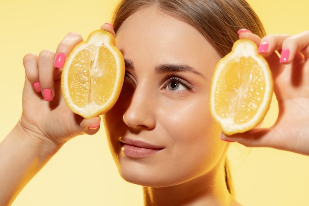 Primo piano di bella giovane donna con fette di limone su sfondo giallo. Concetto di cosmetici, trucco, trattamento naturale ed ecologico, cura della pelle. Pelle lucida e sana, moda, assistenza sanitaria.