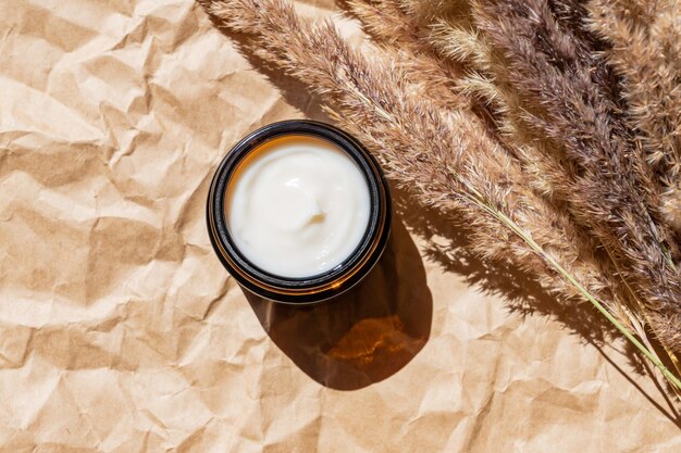 Primo piano di crema al collagene in barattolo di vetro e canne di fiori secchi su fondo beige prodotti di bellezza per la cura della pelle e del corpo