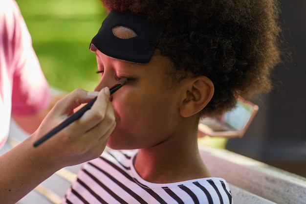 Primo piano di donna irriconoscibile che mette trucco o vernice per il viso sul ragazzo afro-americano che indossa il costume di Halloween