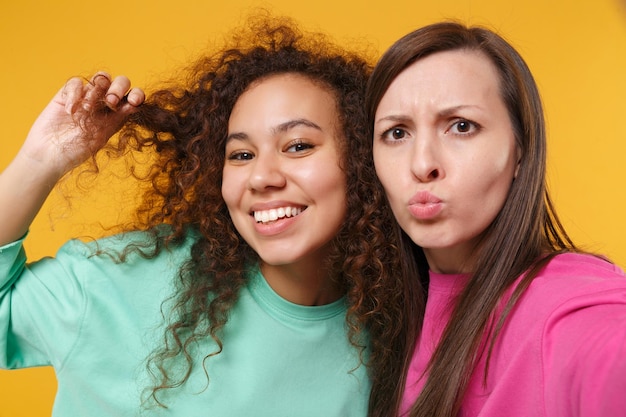 Primo piano di due donne amiche afroamericane europee in abiti rosa verdi in posa isolata su sfondo giallo. Concetto di stile di vita delle persone. Mock up spazio di copia. Facendo selfie sul cellulare.