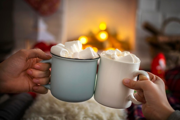 Primo piano di due mani con una tazza di cioccolata calda con marshmallow davanti al caminetto. Godendo la stagione delle vacanze di Natale, momenti felici a casa.