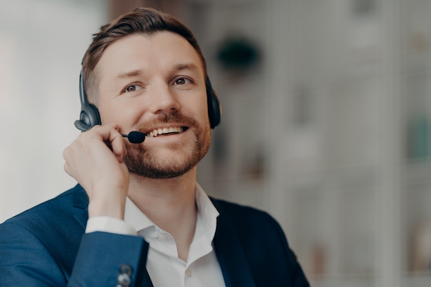 Primo piano di felice impiegato di ufficio bel maschio operatore di call center che indossa l'auricolare e lavora in ufficio, parlando con il cliente online con espressione positiva del viso, messa a fuoco selettiva. Lavoro e occupazione