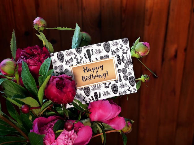 Primo piano di fiori di peonie rosa e vaso su fondo di legno con carta di buon compleanno La foto è stata scattata con luce naturale