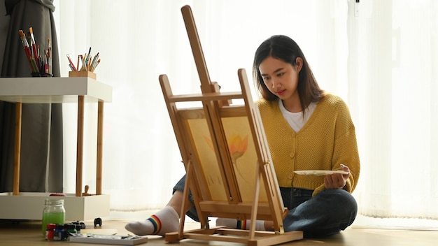 Primo piano di giovane artista femminile seduto sul pavimento tenendo il tubo di vernice e mescolando i colori