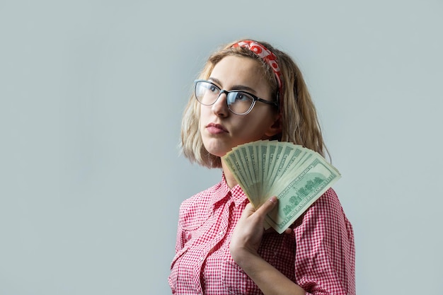 Primo piano di giovane bella donna in una camicia a quadri rossa in bicchieri con dollari americani soldi in mano su sfondo grigio