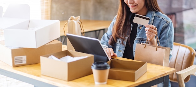 Primo piano di giovane donna asiatica che per mezzo del pc della compressa e della carta di credito per acquisto online con il contenitore di pacchi postale e sacchetti della spesa sul tavolo