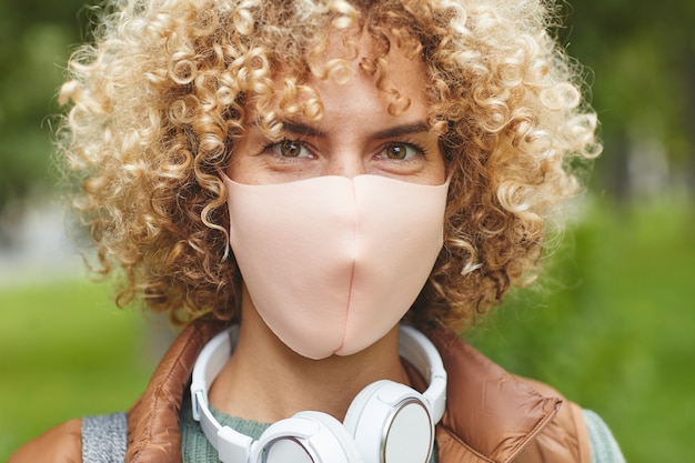 Primo piano di giovane donna con capelli ricci biondi in maschera protettiva che guarda l'obbiettivo in piedi all'aperto