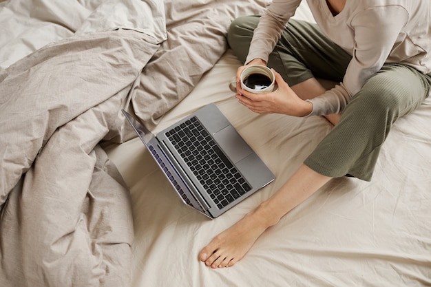 Primo piano di giovane donna seduta nel letto a bere caffè e guardare qualcosa sul computer portatile a casa