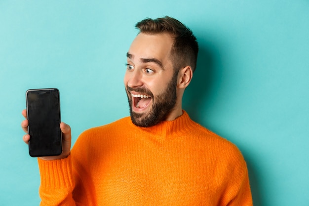 Primo piano di giovane uomo barbuto che mostra lo schermo del telefono e guardando stupito, indossa un maglione arancione