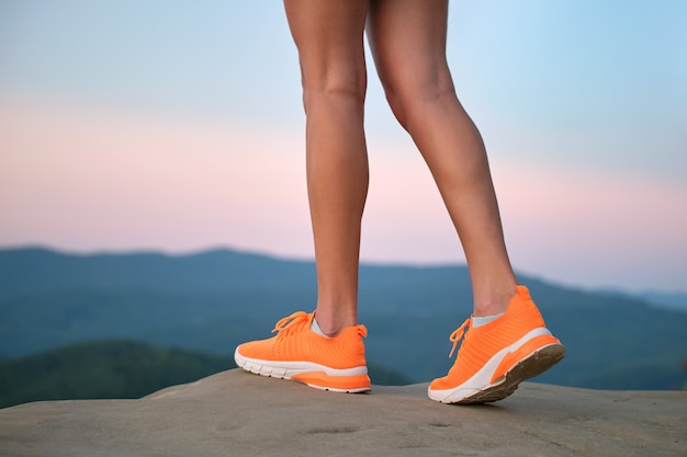 Primo piano di giovani gambe sottili femminili in scarpe da ginnastica arancione brillante in piedi sul sentiero di montagna in sera d'estate. Stile di vita attivo, jogging nella natura ed esercizio sul concetto di aria fresca.