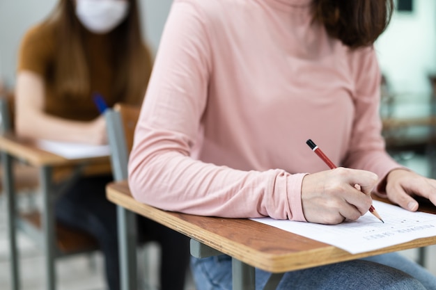 Primo piano di giovani studentesse universitarie si concentrano sul fare l'esame in classe. Una studentessa scrive la risposta degli esami sul foglio delle risposte in classe.