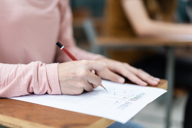 Primo piano di giovani studentesse universitarie si concentrano sul fare l'esame in classe. Una studentessa scrive la risposta degli esami sul foglio delle risposte in classe.