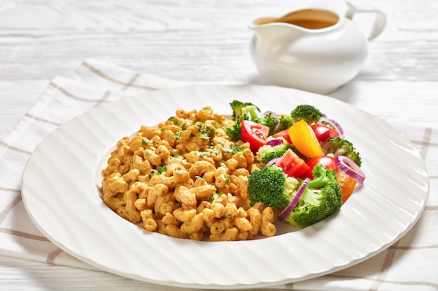 Primo piano di mac vegano e formaggio con salsa di lievito alimentare e insalata di verdure su un piatto bianco su un tavolo di legno bianco con posate d'argento, vista del paesaggio dall'alto