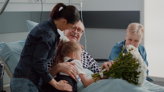 Primo piano di madre e bambino che sorprendono il nonno con la visita nel reparto ospedaliero, portando fiori a un uomo anziano malato. Famiglia in visita al paziente anziano a letto, dando supporto. Colpo a mano libera