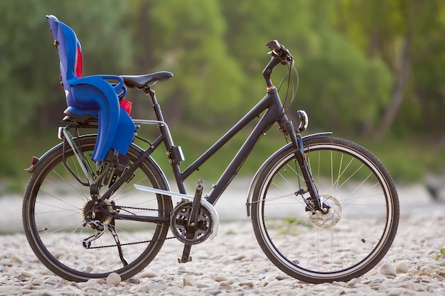 Primo piano di nuova bicicletta nera moderna con il seggiolino per bambini di plastica blu che sta sul supporto laterale acceso dai ciottoli del sole sul fondo verde vago del bokeh di estate degli alberi. Concetto di turismo e viaggi in famiglia.