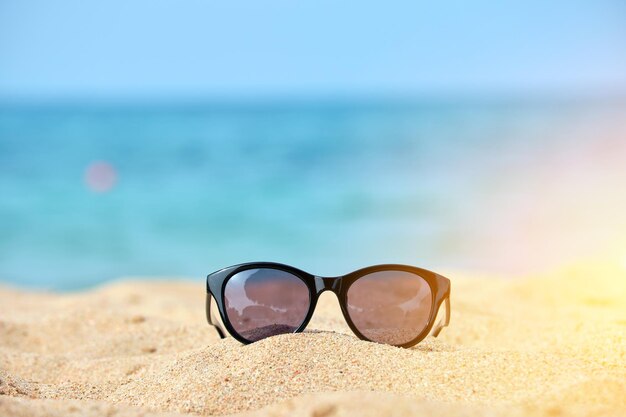 Primo piano di occhiali da sole protettivi neri sulla spiaggia sabbiosa al mare tropicale in una calda giornata di sole Concetto di vacanza estiva