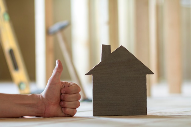 Primo piano di piccola casa di modello di legno e la mano dell'uomo con il gesto del pollice in su