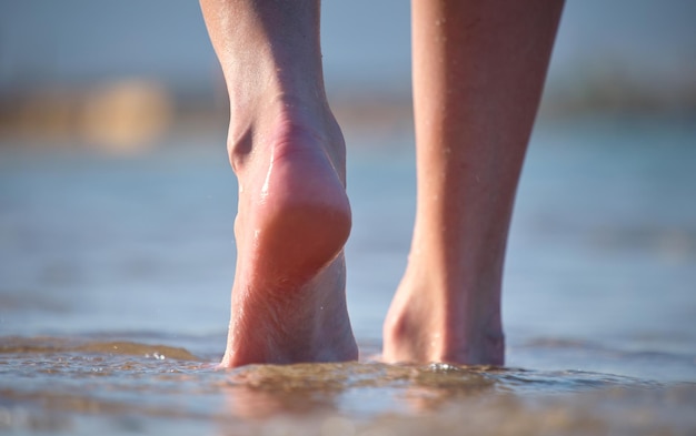 Primo piano di piedi femminili che camminano a piedi nudi sulla sabbia bianca granulosa della spiaggia dorata sullo sfondo dell'acqua blu dell'oceano