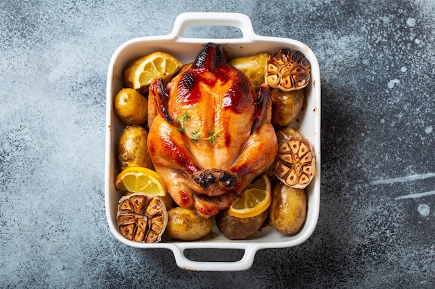 Primo piano di pollo organico croccante succoso intero al forno o arrosto con patate, aglio, limone in casseruola bianca su sfondo rustico in cemento, vista dall'alto